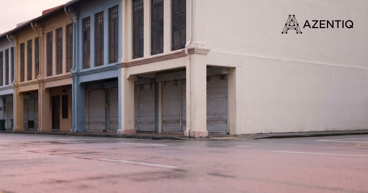 A row of Singapore shophouses at dawn with every shopfront closed behind its roller shutter, and a plain painted gable wall to the right